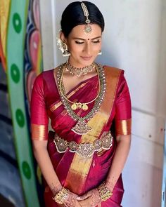 a woman in a red and gold sari with jewelry on her neck, standing next to a colorful wall