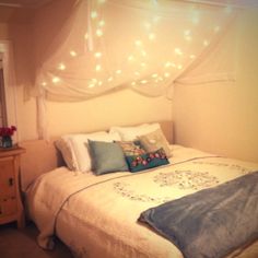 a white bed topped with lots of pillows next to a night stand and window covered in lights