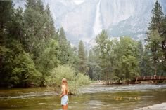 a woman standing in the middle of a river next to a forest filled with trees