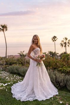 a beautiful woman in a wedding dress standing on the grass with flowers and palm trees behind her