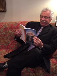 a man sitting on top of a couch holding a book in his hand and smiling at the camera