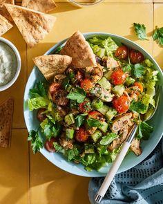 a salad with chicken, lettuce and tomatoes in a bowl next to pita chips