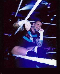 a man sitting on top of a rope in a wrestling ring with his hand up