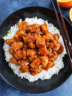 a black plate topped with chicken and rice next to oranges, chopsticks and an orange