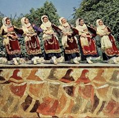 several women in traditional dress are standing on a wall with painted images and trees behind them