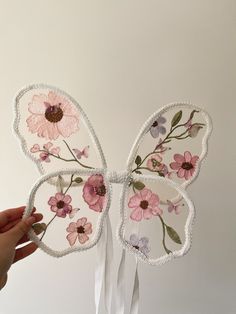 a hand holding a butterfly shaped decoration with pink flowers on it's wings and white ribbon