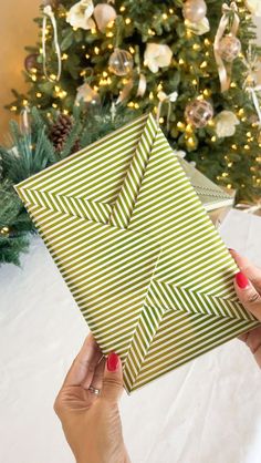 a woman is holding up a green and white gift box in front of a christmas tree