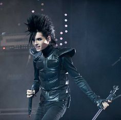 a man with black hair and leather outfit holding a microphone in front of a stage