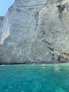 two boats floating in the ocean near a rocky cliff face with blue water and clear sky