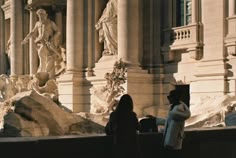 two people standing in front of a building with statues on the side and columns behind them