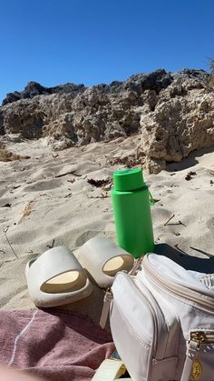 a green cup sitting on top of a sandy beach next to a white bag and sandals