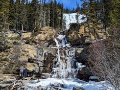 two people standing in front of a waterfall with snow on the ground and trees around