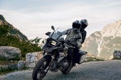 a man riding on the back of a motorcycle down a cobblestone road with mountains in the background