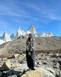 a woman standing on top of a rock covered ground next to snow capped mountain peaks