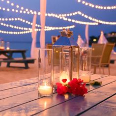 candles and flowers sit on an outdoor table with lights strung over the ocean in the background