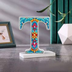 the letter t is decorated with multicolored beads and stands on a wooden table