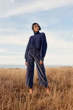 a woman standing in tall grass wearing a blue sweater and wide legged pants with her hands on her hips