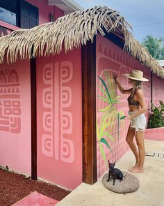 a woman standing next to a pink building with a cat on the ground in front of it