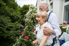 an older couple embracing each other in front of some flowers and bushes on a sunny day