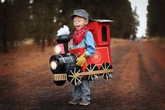 a young boy dressed up as a train with a hat and scarf on, standing in the middle of a dirt road