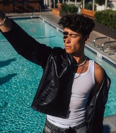 a young man standing in front of a swimming pool wearing a leather jacket and jeans