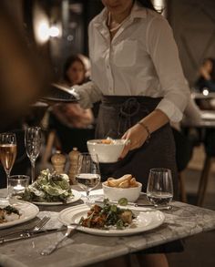 a woman standing at a table filled with food and wine glasses on top of it