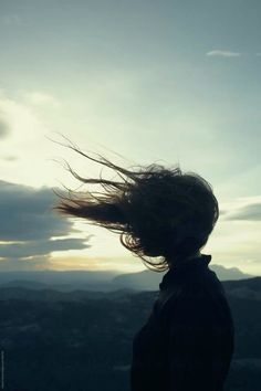 a woman with her hair blowing in the wind on top of a mountain at sunset