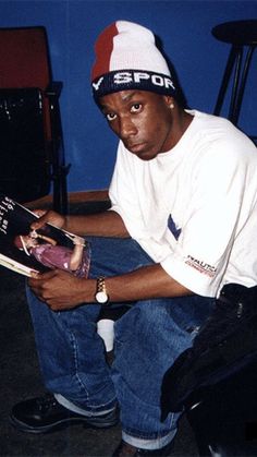 a man sitting down reading a book with his hands on his knees and looking at the camera