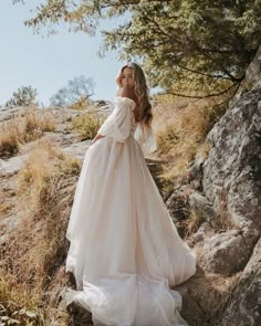 a woman in a wedding dress standing on the side of a mountain with her arms behind her back