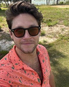 a man wearing sunglasses and an orange shirt sitting in the grass with trees behind him