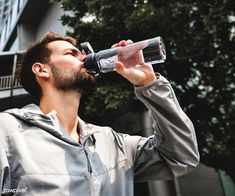 a man drinking from a water bottle while standing in front of a building with trees