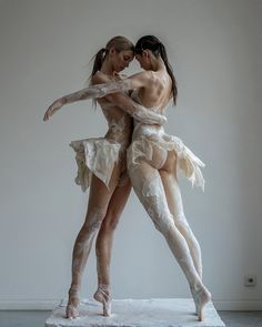 two women in white bodysuits are hugging each other on a marble pedestal with one woman's leg wrapped around the other