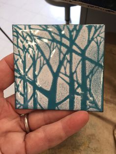 a hand holding up a small square tile with trees painted on it in blue and white