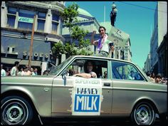 a man riding in the back of a silver car with a sign that says harvey milk