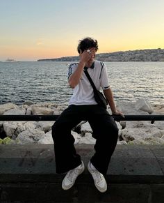 a man sitting on a ledge next to the ocean while talking on a cell phone