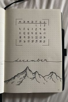 an open notebook with a calendar and mountains drawn on the pages in black ink, sitting on a bed