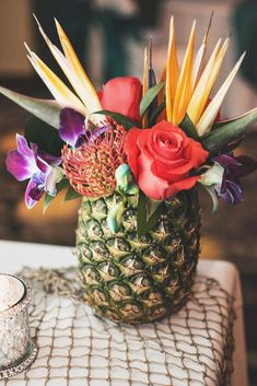 a pineapple vase filled with colorful flowers on top of a white table cloth next to a candle