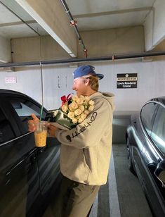 a young man holding flowers next to a car in a parking garage with a drink