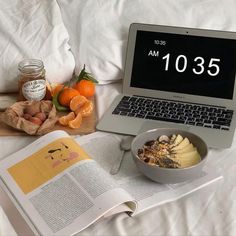 an open book and bowl of food on top of a bed next to a laptop