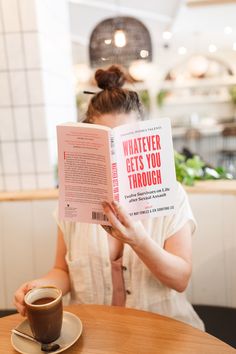 a woman sitting at a table reading a book with a cup of coffee in front of her