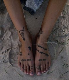 two people with tattoos on their feet standing in the sand near grass and barbed wire