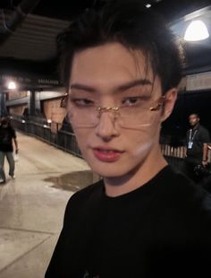 a young man wearing glasses standing in front of a train platform at night with people walking by