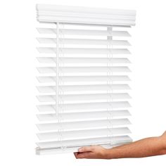 a hand holding a white window blind in front of a white background with the blinds closed