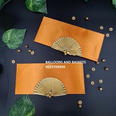 two gold fan shaped napkins sitting on top of a table next to green leaves