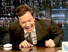 a man laughing while sitting at a table in front of a coffee cup and mug