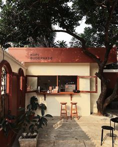 the outside of a small building with tables and stools in front of it, next to a tree