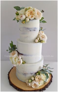 a three tiered cake with white flowers and greenery on the top is sitting on a wood slice