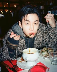 a man eating noodles with chopsticks at a table in a restaurant, while holding a fork above his mouth