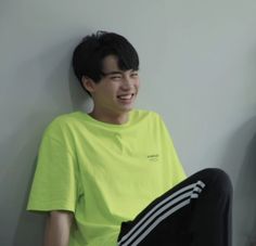 a young man sitting against a wall wearing a yellow shirt and black pants with his legs crossed