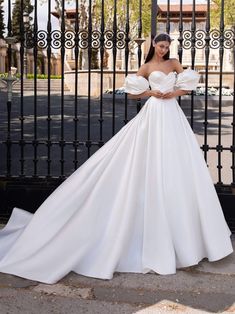 a woman standing in front of a black gate wearing a white wedding dress with puffy sleeves